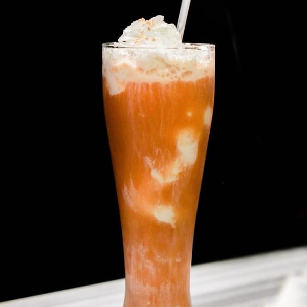 Tall glass of Thai iced tea with whipped cream and a straw against a dark background.